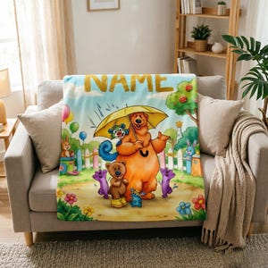 May include: A soft fleece blanket featuring a colorful cartoon illustration of a large brown bear holding a yellow umbrella, surrounded by smaller animal characters, flowers, and a white picket fence, with the word NAME printed at the top.