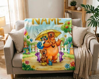May include: A soft fleece blanket featuring a colorful cartoon illustration of a large brown bear holding a yellow umbrella, surrounded by smaller animal characters, flowers, and a white picket fence, with the word NAME printed at the top.