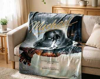 May include: A plush blanket featuring a movie poster design with the name "Michael" in gold script. The image includes a figure in a hat, and the text "In Theaters and May April 24". The blanket is draped over a beige sofa.