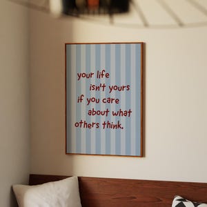 Peut inclure: Une affiche encadrée avec un fond à rayures bleues et blanches et le texte "your life isn't yours if you care about what others think." L'affiche est dans une pièce avec une table en bois, un oreiller et une assiette avec des oranges.