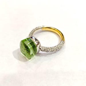 May include: A gold and silver ring with a large, faceted green gemstone. The band is gold on the inside and silver with small, clear stones on the outside. The green stone is the focal point of the ring.