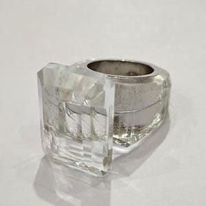 May include: A clear, faceted crystal ring with a silver band. The ring features a large, rectangular crystal element and a polished silver band. The design is modern and geometric, suitable for fashion jewelry.