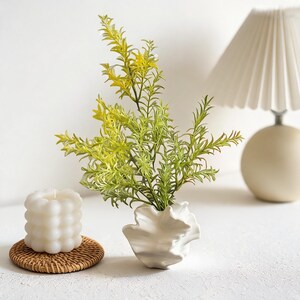A white ceramic vase with an organic, shell-like wavy design, holding vibrant green leafy branches against a white background.