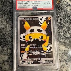 May include: A graded Pok&eacute;mon card featuring a Pikachu character in a black and yellow skull-themed design. The card is encased in a protective holder and includes text in Japanese and English, with a GEM MT 10 grade.