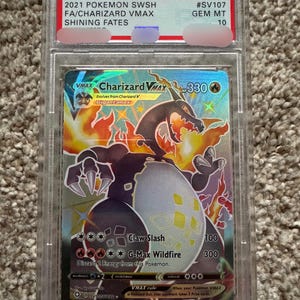 Puede incluir: Una carta Pokémon graduada con Charizard VMAX del set Shining Fates. La carta muestra una vibrante ilustración de Charizard con acentos de fuego y el texto "Claw Slash" y "G-Max Wildfire". La carta está dentro de un soporte protector.