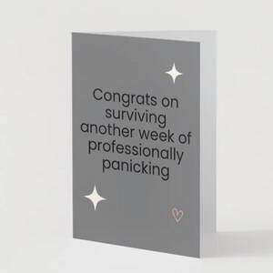 May include: A folded greeting card with a grey front. The card reads "Congrats on surviving another week of professionally panicking" in black text. Two white starbursts and a small pink heart are also on the front.