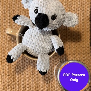 Puede incluir: Un koala de ganchillo gris claro hecho a mano con detalles negros en la nariz, las patas y los pies. El koala está sentado en una taza beige. Un círculo morado con el texto "PDF Pattern Only" está en la esquina inferior derecha.