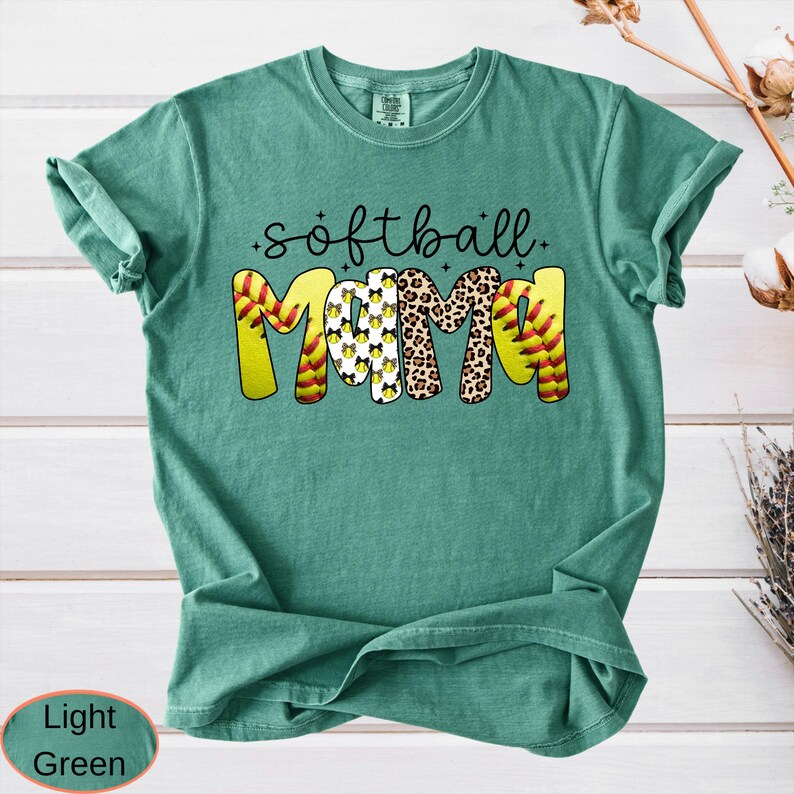 May include: Light green t-shirt with the words "softball mama" in a playful design. The word "mama" is decorated with a softball, leopard print, and softball patterns. The shirt is a comfortable, casual style.