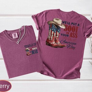 May include: Two berry-colored t-shirts. One shirt has a small graphic of a cowboy boot with the text "BROUGHT TO YOU COURTESY OF WHITE & BLUE". The other shirt features a graphic of a cowboy boot with the American flag and the text "WE'LL PUT A BOOT IN YOUR ASS The American Way".