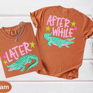 Puede incluir: Dos camisetas marrones con las frases "LATER" y "AFTER WHILE" en letras rosas. Cada camiseta presenta un gr&aacute;fico de caim&aacute;n verde azulado con acentos de estrellas rosas y amarillas. Una cadena de plata est&aacute; sobre una camiseta.