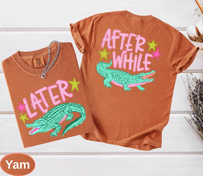 Puede incluir: Dos camisetas marrones con las frases "LATER" y "AFTER WHILE" en letras rosas. Cada camiseta presenta un gr&aacute;fico de caim&aacute;n verde azulado con acentos de estrellas rosas y amarillas. Una cadena de plata est&aacute; sobre una camiseta.