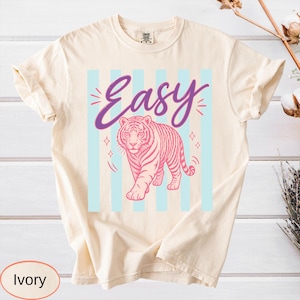 May include: Ivory-colored t-shirt featuring a pink tiger design with the word "Easy" in a purple script. The tiger is set against a backdrop of light blue vertical stripes. The shirt has a classic crew neck and short sleeves.