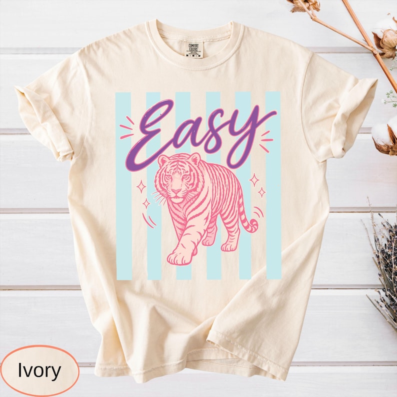 May include: Ivory-colored t-shirt featuring a pink tiger design with the word "Easy" in a purple script. The tiger is set against a backdrop of light blue vertical stripes. The shirt has a classic crew neck and short sleeves.