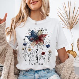 May include: White t-shirt featuring a floral design in red, white, and blue, with the word "America" and the dates "1776 2026" in black script. The design includes butterflies and stars.