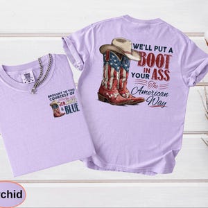 May include: Orchid-colored t-shirts with a cowboy boot graphic. One shirt features a small boot graphic and text, "BROUGHT TO YOU COURTESY OF THE RED, WHITE & BLUE." The other shirt has a larger graphic of a cowboy boot with a hat and the text, "WE'LL PUT A BOOT IN YOUR ASS The American Way."