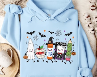 May include: A light blue hooded sweatshirt featuring a graphic print of five cartoon characters dressed for Halloween: a ghost, a mummy apple, a glue stick wearing a witch hat, a vampire notebook, and a pencil, surrounded by bats and spiderwebs.