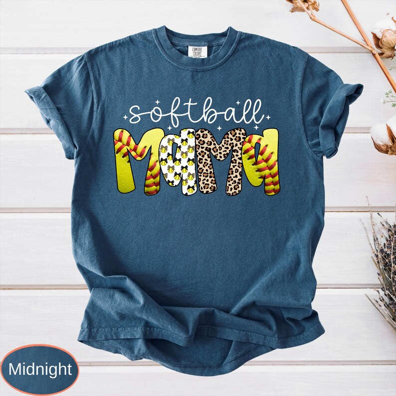 May include: A navy blue t-shirt with the words "softball Mama" in white script and a colorful design. The word "Mama" features a softball design, leopard print, and a pattern of softballs. The shirt is a comfortable, casual style.