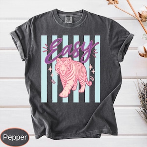 May include: Dark gray t-shirt featuring a pink tiger graphic with the word "Easy" in a cursive font. The design includes light blue vertical stripes and star accents. The shirt has rolled-up sleeves.