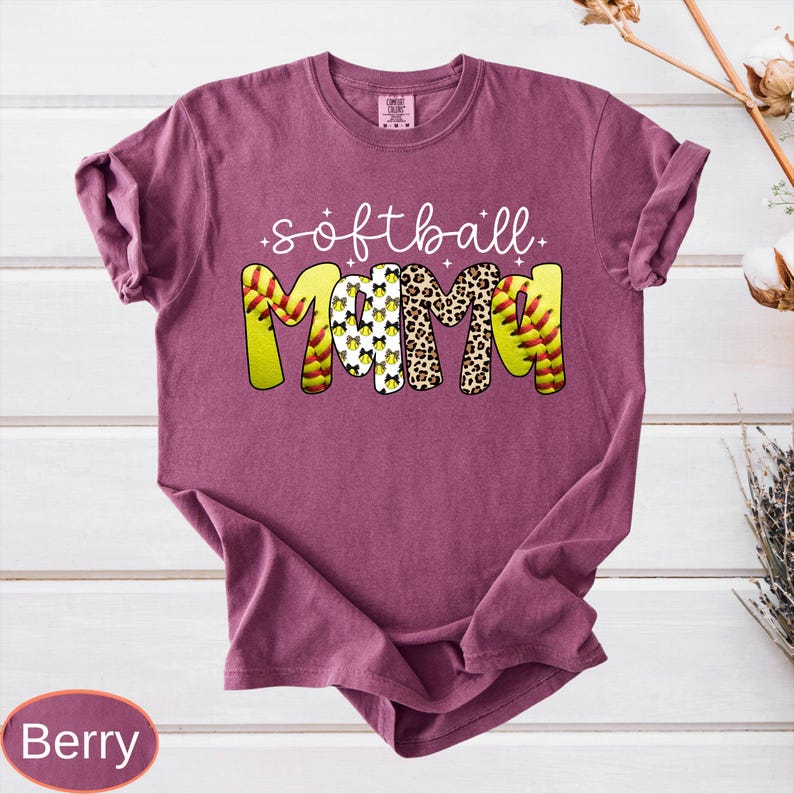 May include: Berry-colored t-shirt with the words "softball Mama" in a playful design. The word "Mama" features a softball pattern, leopard print, and a pattern of softball images. The shirt is a comfortable, casual style.