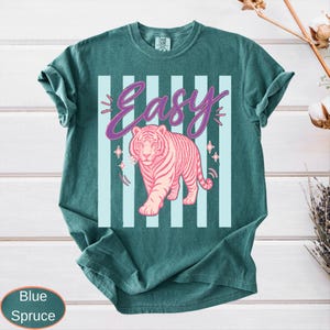 May include: Blue spruce t-shirt with a pink tiger graphic and the word "Easy" in a cursive font. The tiger is walking forward, and the background has light blue vertical stripes. The shirt is a solid color.