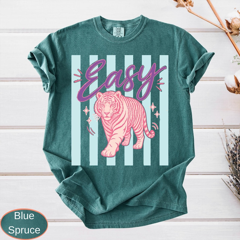 May include: Blue spruce t-shirt with a pink tiger graphic and the word "Easy" in a cursive font. The tiger is walking forward, and the background has light blue vertical stripes. The shirt is a solid color.