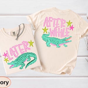 Puede incluir: Dos camisetas color marfil con un dise&ntilde;o de cocodrilo verde azulado. Una camiseta dice "LATER" con una estrella rosa, y la otra dice "AFTER WHILE" con una estrella rosa. El cocodrilo tiene detalles rosas.