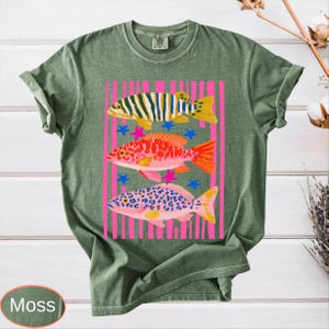 May include: Moss green t-shirt featuring a colorful fish design. The graphic includes three distinct fish with vibrant patterns, set against a pink striped background with blue stars. The shirt is made of a soft, comfortable material.