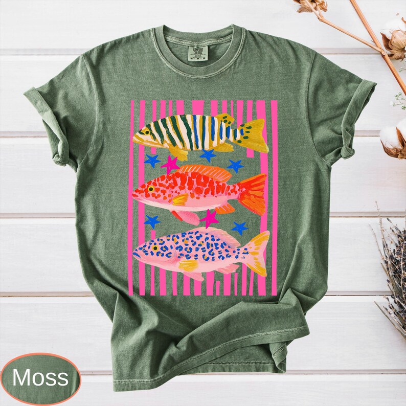 May include: Moss green t-shirt featuring a colorful fish design. The graphic includes three distinct fish with vibrant patterns, set against a pink striped background with blue stars. The shirt is made of a soft, comfortable material.