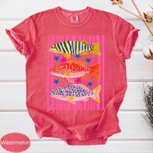 May include: A red t-shirt with a graphic of three colorful fish. The fish are yellow, green, orange, and blue, with pink stars and a pink striped background. The word "Watermelon" is at the bottom.
