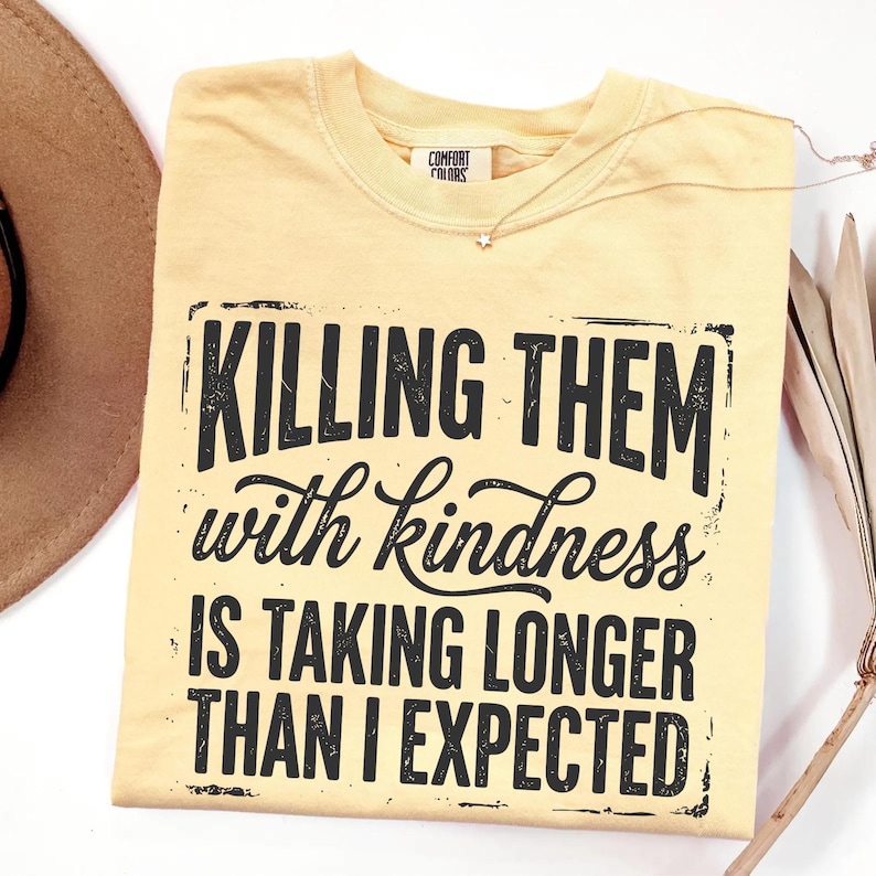 Op de afbeelding: Een lichtgeel t-shirt met korte mouwen en een zwarte grafische opdruk met de tekst "KILLING THEM with kindness IS TAKING LONGER THAN I EXPECTED." Het shirt heeft een ronde hals en een Comfort Colors-merklabel aan de binnenkant van de kraag.