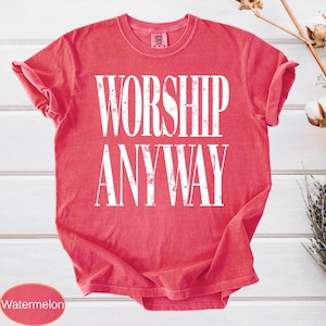 May include: A watermelon-colored t-shirt with the words "WORSHIP ANYWAY" printed in large, white, distressed lettering. The shirt has a crew neck and short sleeves, with the sleeves rolled up. The shirt is made of a soft material.