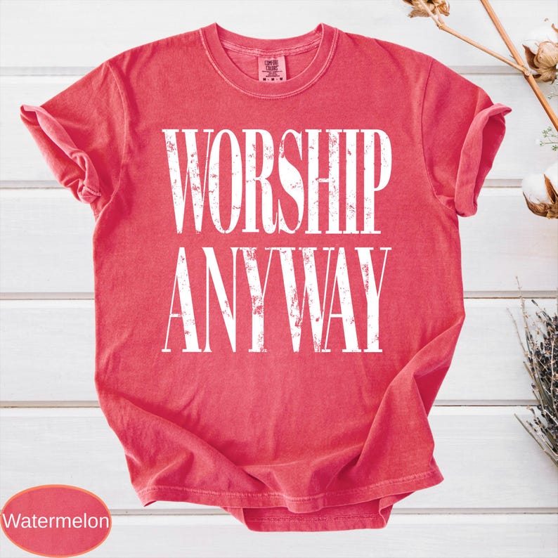 May include: A watermelon-colored t-shirt with the words "WORSHIP ANYWAY" printed in large, white, distressed lettering. The shirt has a crew neck and short sleeves, with the sleeves rolled up. The shirt is made of a soft material.