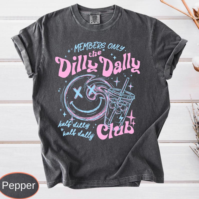 A dark fabric featuring a distressed retro print. The words "MEMBERS ONLY," "the Dilly Dally Club," and "half dilly, half dally" are in a combination of pink and blue bubbly script. A central blue swirl smiley face with X-eyes is next to a blue-line iced coffee cup. The entire graphic has an authentic aged, screen-printed texture, framed by tiny pink and blue starbursts.