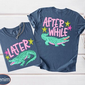 Puede incluir: Dos camisetas de color denim con dise&ntilde;os gr&aacute;ficos. Una camiseta presenta el texto "LATER" con una estrella rosa y un caim&aacute;n verde. La otra camiseta tiene "AFTER WHILE" con un caim&aacute;n verde y estrellas rosas y amarillas.