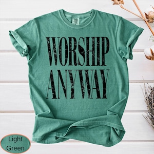May include: A light green t-shirt with the words "WORSHIP ANYWAY" printed in large, distressed black letters. The shirt has a crew neck and rolled-up sleeves. The bottom left corner of the image has a light green oval with the words "Light Green".