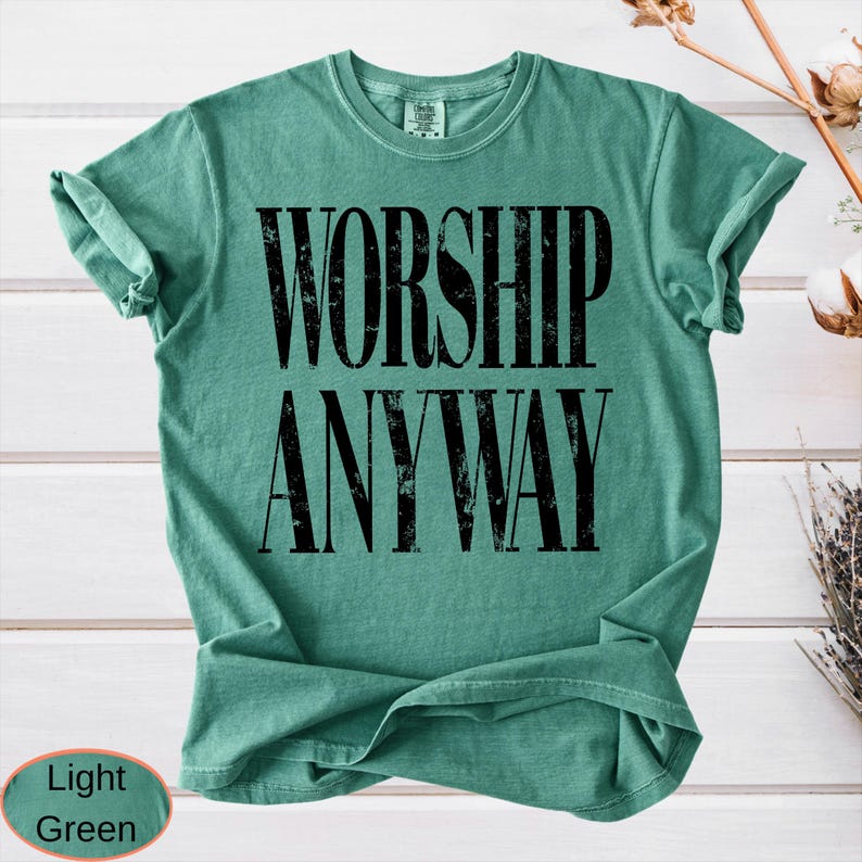 May include: A light green t-shirt with the words "WORSHIP ANYWAY" printed in large, distressed black letters. The shirt has a crew neck and rolled-up sleeves. The bottom left corner of the image has a light green oval with the words "Light Green".