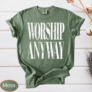 May include: A moss green t-shirt with the words "WORSHIP ANYWAY" printed in large white letters. The shirt has rolled-up sleeves and a crew neck. The word "Moss" is in a small oval at the bottom left.