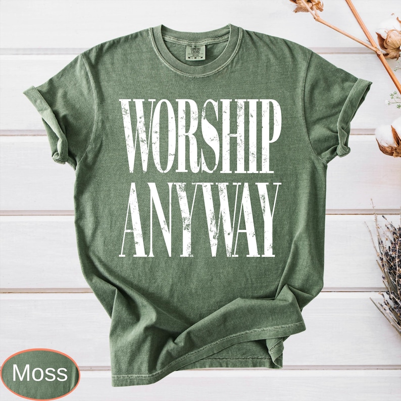 May include: A moss green t-shirt with the words "WORSHIP ANYWAY" printed in large white letters. The shirt has rolled-up sleeves and a crew neck. The word "Moss" is in a small oval at the bottom left.