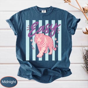May include: Midnight blue t-shirt featuring a pink tiger graphic with the word "Easy" in a cursive font. The design includes light blue vertical stripes and star accents, creating a retro aesthetic.