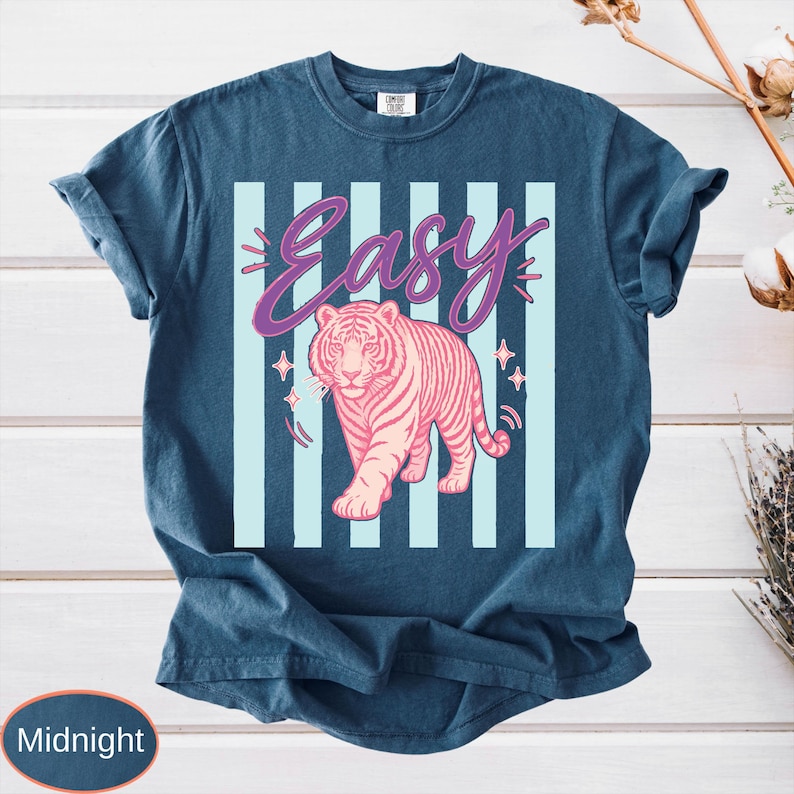 May include: Midnight blue t-shirt featuring a pink tiger graphic with the word "Easy" in a cursive font. The design includes light blue vertical stripes and star accents, creating a retro aesthetic.