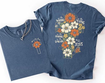 May include: A blue short-sleeve t-shirt featuring a small cross and flower graphic on the front left chest and a large cross filled with orange and white flowers and a butterfly on the back, with the text In the waiting, in the healing, in the hurting, in the searching, there was Jesus.