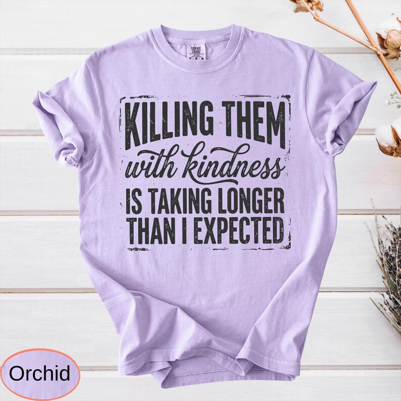 Op de afbeelding: Een lichtpaars t-shirt met korte mouwen en zwarte tekst die luidt: "KILLING THEM with kindness IS TAKING LONGER THAN I EXPECTED." Het shirt heeft een ronde hals en een relaxte pasvorm, met de afbeelding gecentreerd op de borst.