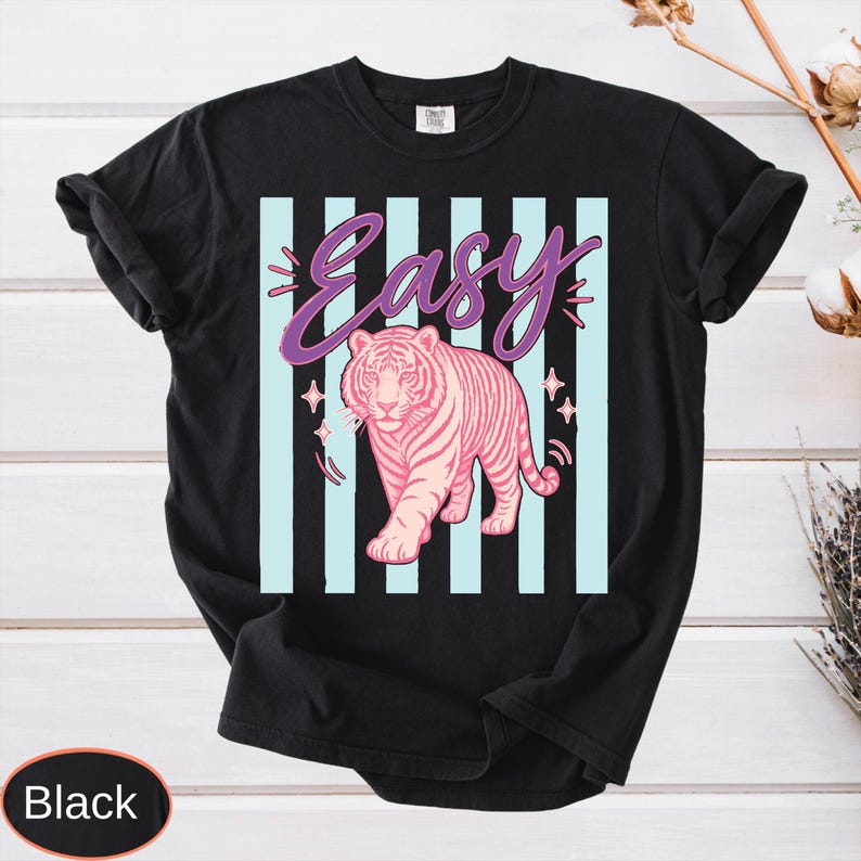 May include: Black t-shirt with a pink tiger graphic and the word "Easy" in purple script. The design includes light blue vertical stripes and star accents, creating a bold and eye-catching design.