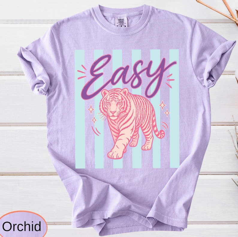 Orchid-colored t-shirt with a pink tiger graphic and the word "Easy" in cursive. The tiger is set against a backdrop of light blue vertical stripes and sparkling star accents. The shirt is made of a soft material.