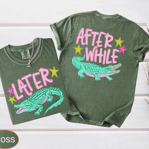 Puede incluir: Dos camisetas verde musgo con gr&aacute;ficos de caimanes. Una camiseta tiene "LATER" y la otra "AFTER WHILE" en texto rosa con acentos de estrellas amarillas y rosas. Una cadena de plata est&aacute; en la primera camiseta.