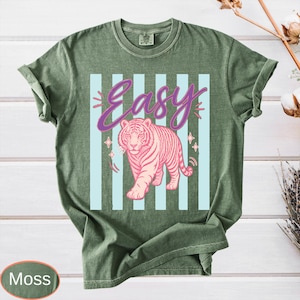 May include: Moss green t-shirt with a pink tiger graphic and the word "Easy" in a script font. The tiger is centered on light blue vertical stripes. The shirt has short sleeves and a crew neck.