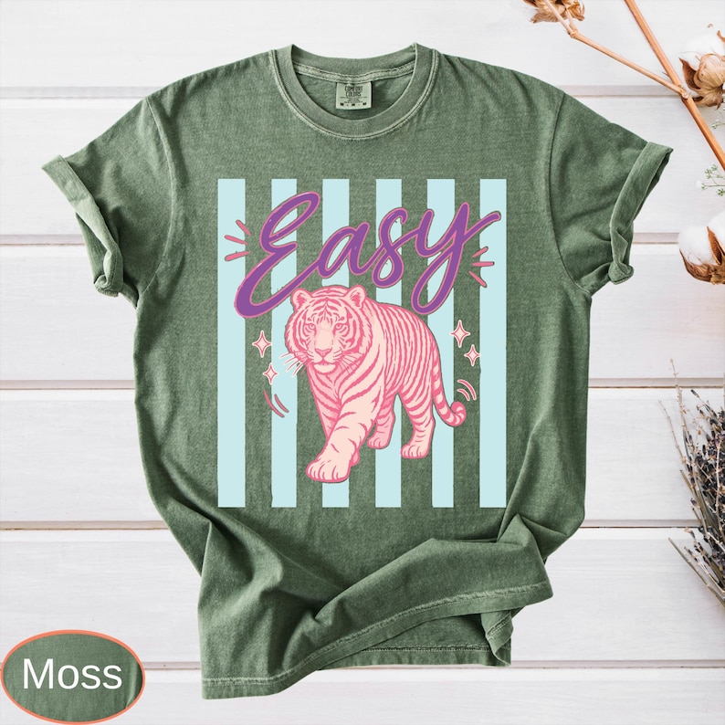 May include: Moss green t-shirt with a pink tiger graphic and the word "Easy" in a script font. The tiger is centered on light blue vertical stripes. The shirt has short sleeves and a crew neck.