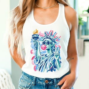 May include: White tank top featuring a graphic of the Statue of Liberty wearing sunglasses and blowing a bubblegum bubble. The Statue of Liberty is holding a torch and making a peace sign. The design has blue, pink, and yellow accents.