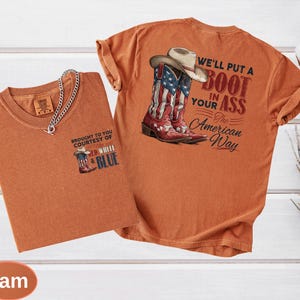 May include: Two orange-colored t-shirts. One shirt features a graphic of a cowboy boot with the American flag design and the text "BROUGHT TO YOU COURTESY OF RED, WHITE & BLUE". The other shirt has a cowboy boot with the American flag design and the text "WE'LL PUT A BOOT IN YOUR ASS The American Way".