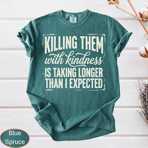 Op de afbeelding: Een blauwspar-kleurig t-shirt met korte mouwen en de gebroken witte tekst "KILLING THEM with kindness IS TAKING LONGER THAN I EXPECTED" gedrukt in een combinatie van vetgedrukte blokletters en een cursief lettertype op de borst.